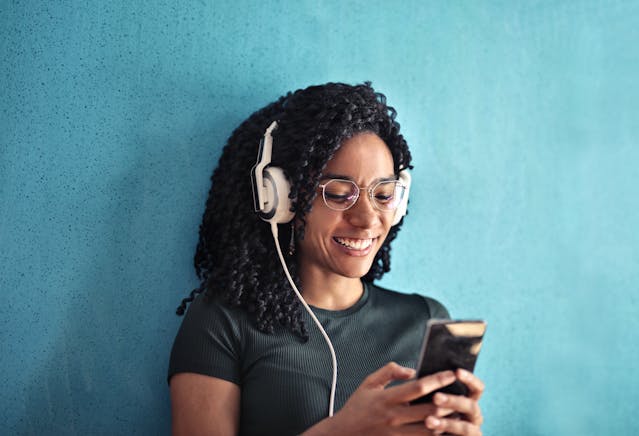 A smiling woman wearing headphones while browsing her phone.