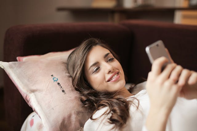 A smiling woman lying on a couch while typing messages on her phone.