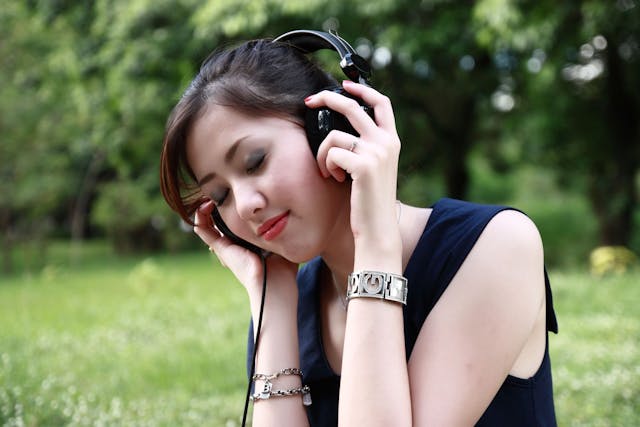 両手でヘッドホンを持ち、目を閉じて音楽を聴いている女性。