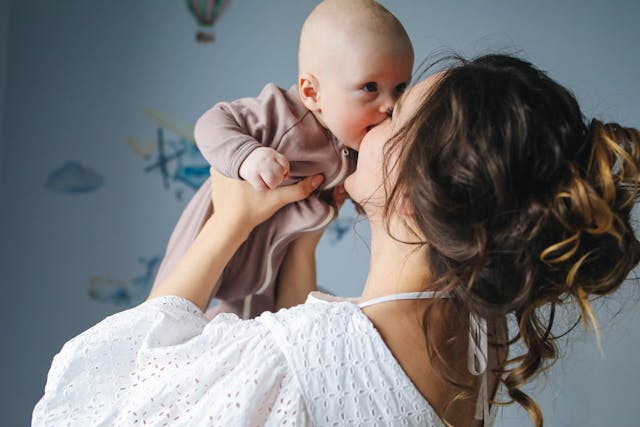 A mom with a messy bun lifting her baby in the air while kissing him.