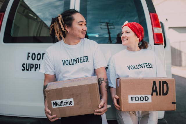 Twee vrijwilligers met dozen met het label "medicijnen" en "voedsel" kijken elkaar glimlachend aan.