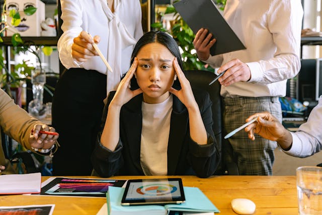オフィスで複数の同僚が話しかけようとする中、こめかみをマッサージするストレス顔の女性。