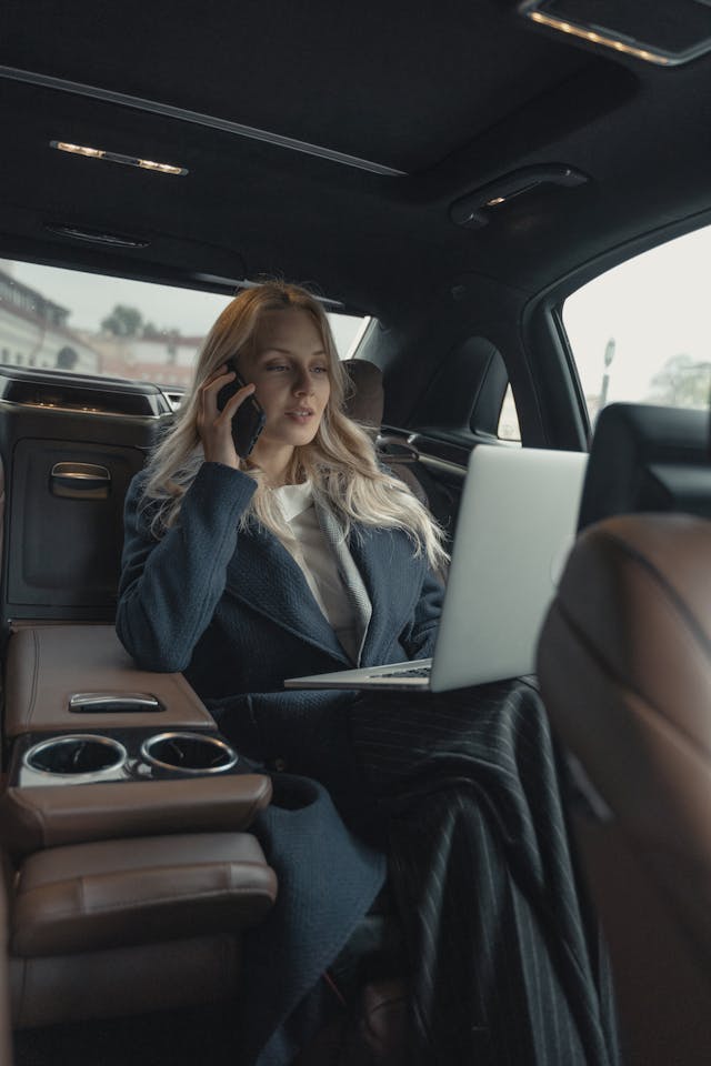 A mom wearing a business suit taking a phone call while typing on her laptop in the back of a car.