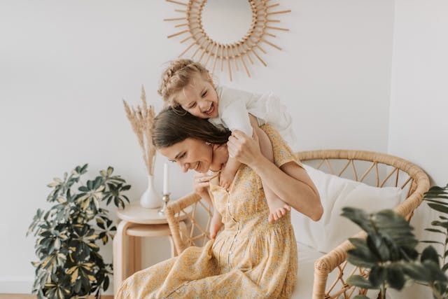 Uma mãe com um vestido floral sentada enquanto carrega a sua filha pequena e sorridente às costas.