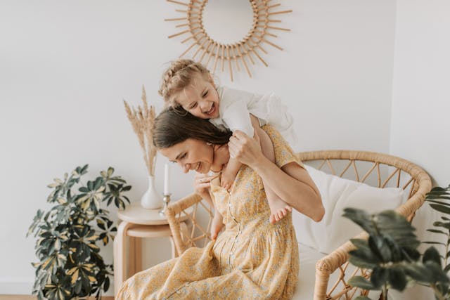 Une maman avec une robe à fleurs, assise, portant sur son dos sa jeune fille souriante.