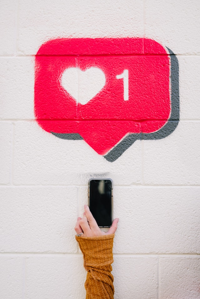 karsten-winegeart-60GsdOMRFGc-unsplash | Path Social Una persona che tiene in mano uno smartphone contro un muro bianco con un graffito dell'icona "like" rosa Instagram .