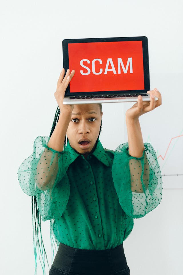 pexels-n-voitkevich-7172786 | Path Social A woman in a green blouse holding up a laptop with the word “SCAM” displayed against a red background.