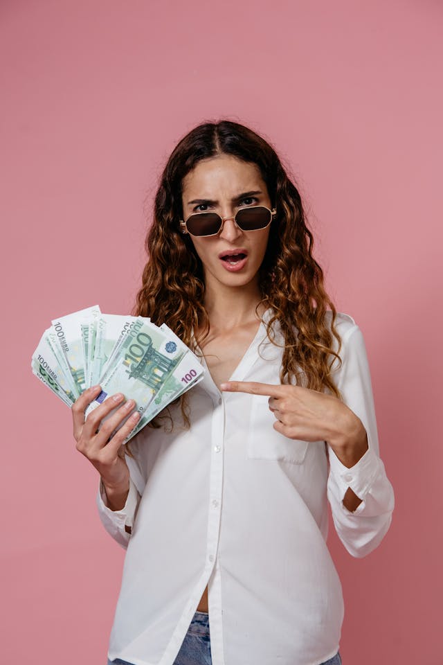 pexels-pavel-danilyuk-8638317 | Path Social A woman in a white long-sleeved shirt and sunglasses pointing at a fan of cash bills she’s holding.