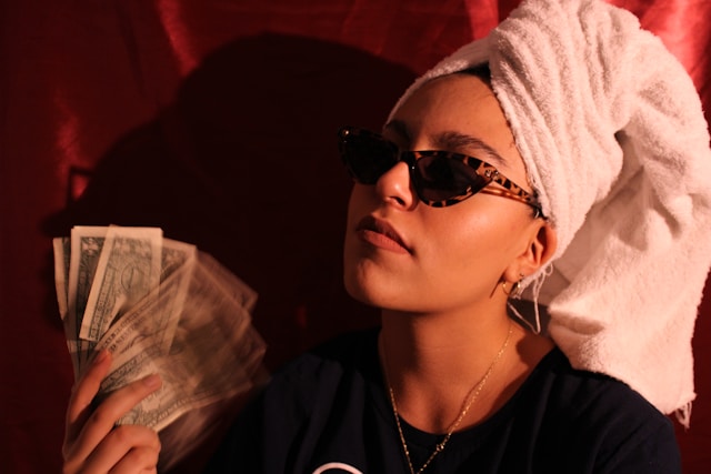 vanessa-murrieta-ZATo07FA5v8-unsplash | Path Social A smug-looking woman with sunglasses and a towel on her head fanning herself with dollar bills.