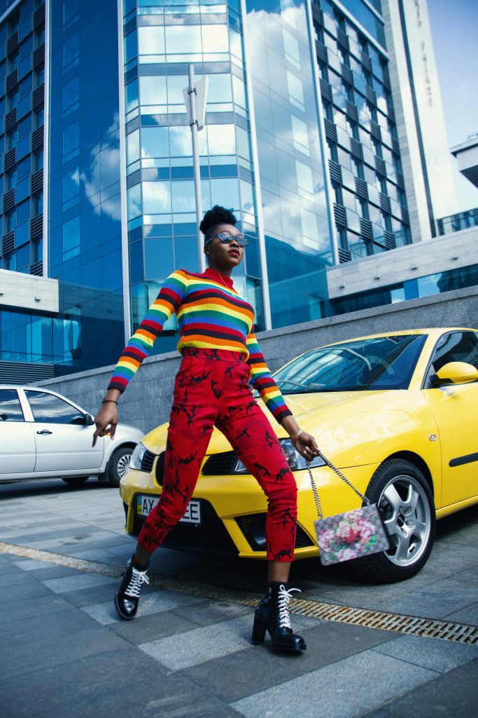 pexels-godisable-jacob-226636-1075776 | Path Social Eine Modeaufnahme eines Models, das einen Regenbogenpullover, rote Jeans und eine geblümte Handtasche trägt, während es die Straße überquert.