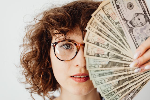pexels-karolina-grabowska-7680625 | Path Social Una mujer de pelo rizado que cubre la mitad de su cara con un abanico de billetes de dólar.