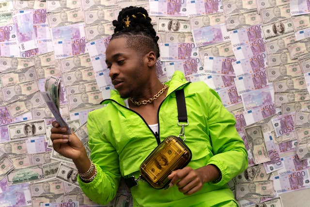 pexels-cottonbro-5911973 | Path Social Um homem com um casaco verde néon deitado num chão cheio de dinheiro enquanto segura notas de dinheiro.