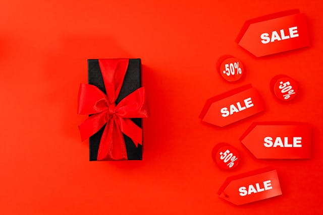 pexels-karolina-grabowska-5625046 | Path Social Regalo con papel de regalo negro y un lazo rojo sobre una superficie roja con carteles de oferta y 50% de descuento a la derecha.