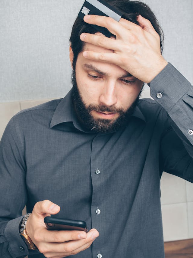 Um homem de aspeto stressado, com uma camisa de botões cinzenta, a olhar para o telemóvel enquanto tem uma mão na testa.