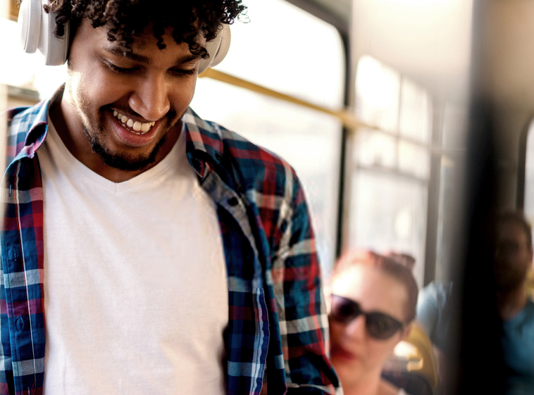 1png | Path Social Lächelnder Mann mit Kopfhörern und kariertem Hemd, der während einer Fahrt mit öffentlichen Verkehrsmitteln auf sein Telefon schaut.