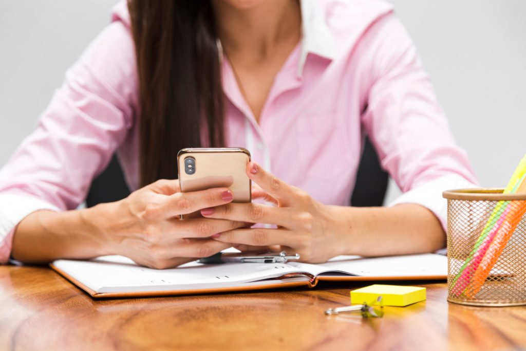 A person sitting at a desk using a smartphone with an open notebook and stationery.